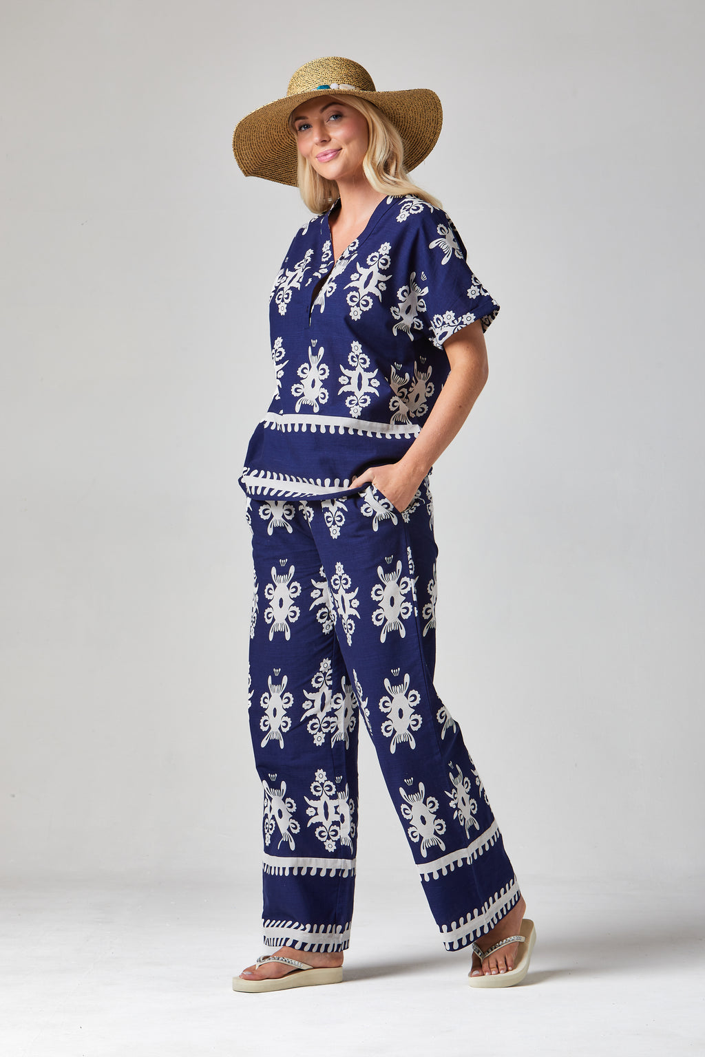 Buy Online Athena Trousers Alcie Collins - Woman wearing a blue and white patterned outfit with a wide-brimmed hat on a plain background