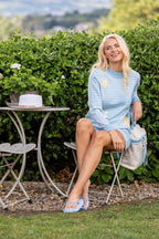 Buy Online Celine Shorts Alice Collins - Woman sitting outdoors in a garden setting, wearing a light blue dress with sunflower patterns.
