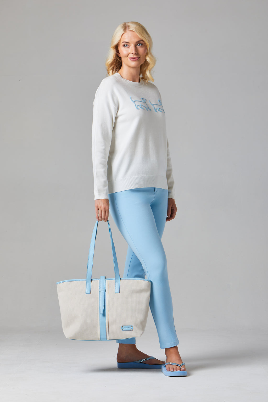 Buy Online Jessie & Pippa Pullover Alice Collins -  Woman wearing a light gray sweater and blue pants, holding a matching tote bag on a gray background