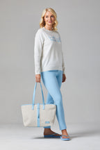 Buy Online Jessie & Pippa Pullover Alice Collins -  Woman wearing a light gray sweater and blue pants, holding a matching tote bag on a gray background