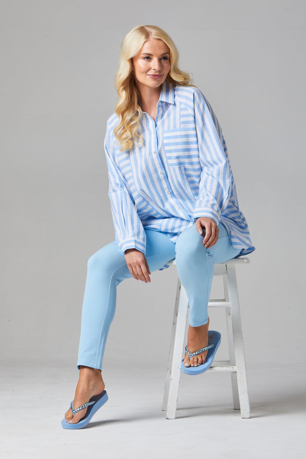 Buy Online Alison Shirt - Alice Collins  Woman sitting on a stool wearing a blue and white striped shirt and light blue jeans against a gray background.